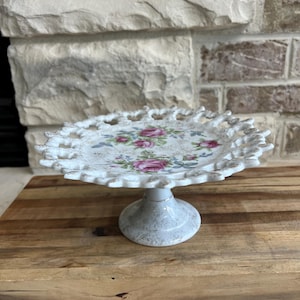 May include: A white ceramic cake stand with a scalloped edge and a floral design featuring pink roses. The stand has a pedestal base and is displayed on a wooden surface. The stand is approximately 8 inches in diameter.
