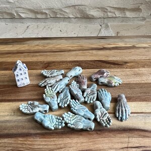 May include: A collection of 15 ceramic hand-shaped pendants in shades of blue, green, and brown. The pendants are arranged on a wooden surface.