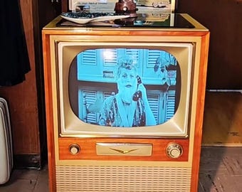 May include: Vintage television with a wooden cabinet and a screen displaying a black and white image. The TV has a light-colored speaker grill and a round dial. Above the TV are framed vintage advertisements. The text "Rag Doll Betty's Vintage" is at the bottom.