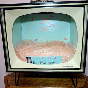 May include: A vintage-style display case designed to resemble an old television set. The case has a black frame with a cream-colored interior and a light blue backdrop with retro starburst and arrow designs. The display contains a peach-colored fabric. The text "Rag Doll Betty's" is visible.