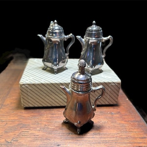 Pode incluir: Três pequenas bules prateados com tampas e alças. Dois estão em uma caixa listrada, e um está na frente. Os bules têm um design clássico com um bico e três pés pequenos. O fundo é preto.