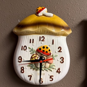 May include: A ceramic wall clock shaped like a mushroom with a yellow cap and white stem. The clock face features brown numbers from 1 to 12, a central illustration of two smaller mushrooms, and black hour and minute hands with a red second hand.