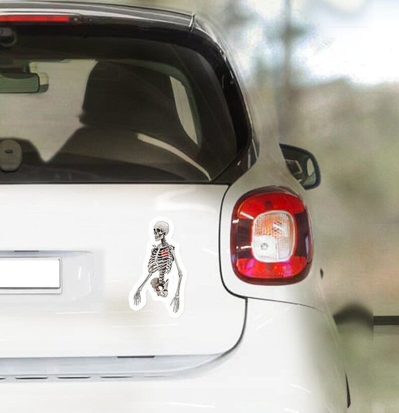 Peut inclure: Une voiture blanche avec un autocollant de squelette sur la vitre arri&egrave;re. Le squelette est debout, les bras tendus. Le squelette a un c&oelig;ur rouge dans la poitrine.