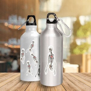 Peut inclure: Deux bouteilles d'eau argent&eacute;es avec un motif de squelette. La bouteille de gauche a plusieurs squelettes, tandis que la bouteille de droite a un grand squelette.