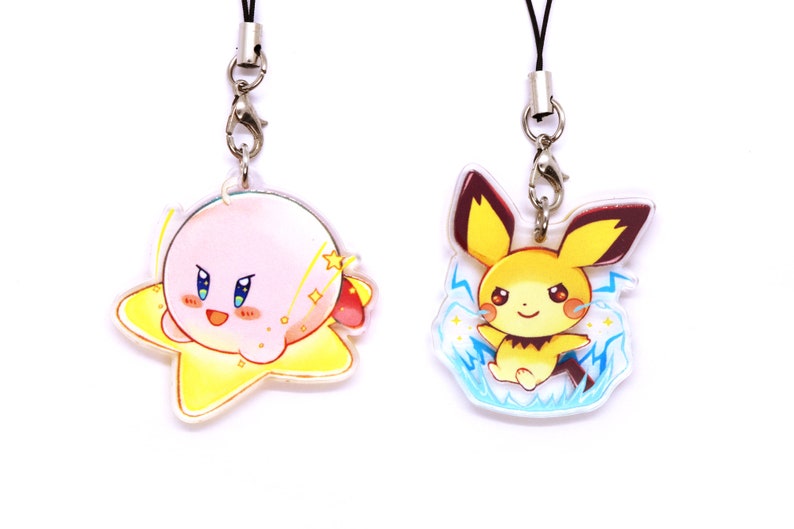 May include: Two colorful character charms on black lanyards with silver clasps. The charm on the left is pink and yellow and features a cartoon character with a star shape. The charm on the right is yellow and blue and features a cartoon character with lightning bolts.