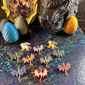 May include: A collection of dragon and griffin enamel pins in various colors, including red, blue, and gold, displayed on a shimmering surface. The image also features dragon eggs and a framed illustration with a gold frame.
