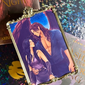 Può includere: Una stampa rettangolare incorniciata color oro con un'illustrazione romantica di un uomo con le ali e una donna. L'uomo ha lunghi capelli castani e indossa una giacca blu. La donna ha lunghi capelli neri e indossa un abito viola.