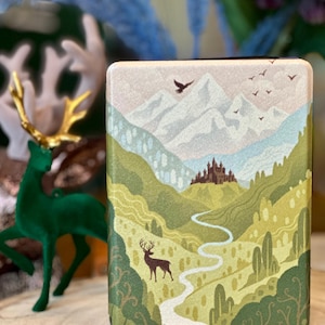 May include: A rectangular box featuring a colourful illustration of a mountain range, a castle, a winding path, and a deer. The sky is light blue with white clouds and birds in flight. The mountains are white with a pale pink hue. The path is light green and white. The deer is brown with antlers. The castle is grey with white towers. The box is made from a robust material and has a smooth finish.