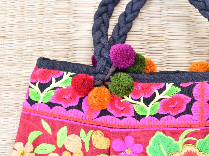 May include: A red and black embroidered tote bag with colorful pom-poms attached to the handles. The bag has a floral design with pink, green, yellow, and orange flowers.