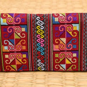 May include: A rectangular, embroidered clutch with a vibrant, geometric pattern in red, yellow, blue, and pink. The clutch has a black zipper and a green pom-pom tassel. The background is a woven mat.
