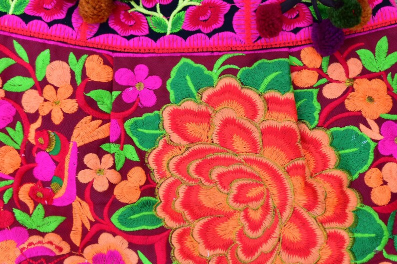 May include: Close-up of a vibrant embroidered fabric with a large red, orange, and pink flower in the center. The fabric is decorated with a variety of other flowers in shades of pink, green, and orange.