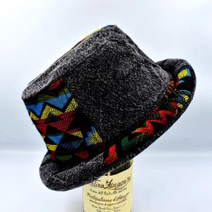 Puede incluir: Un sombrero de copa gris oscuro con un colorido patrón geométrico en el borde y la banda. El sombrero está hecho de una tela texturizada y tiene una forma de sombrero de copa clásica. El patrón colorido incluye zigzags rojos, amarillos, azules y verdes.