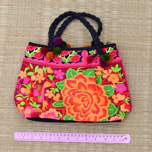 May include: A pink and red embroidered tote bag with black handles and floral designs. The bag has a black trim and pink pom-poms.