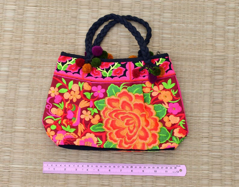 May include: A pink and red embroidered tote bag with black handles and floral designs. The bag has a black trim and pink pom-poms.