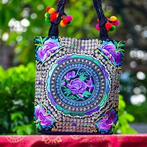 May include: Black tote bag with intricate floral embroidery in purple, blue, and green. The bag features a central circular design with a rose and is accented with colorful pom-poms on the handles. The bag is made of a sturdy material and is suitable for carrying items.