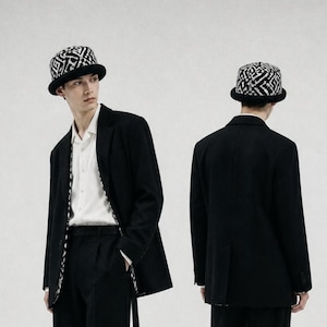 Può includere: Un blazer nero indossato sopra una camicia bianca abbottonata e pantaloni neri, abbinato a un cappello da pescatore con motivo bianco e nero. Il blazer presenta una fodera coordinata con motivo visibile lungo i bordi dell'apertura.