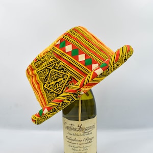Puede incluir: Un sombrero colorido con ala ancha, con diseños geométricos rojos, amarillos, verdes y negros. El sombrero está colocado sobre una botella de vino verde.