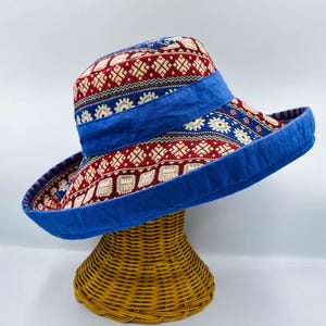 May include: A wide-brimmed hat with a vibrant pattern of red and white geometric shapes on a navy blue background. The hat features a wide, solid blue band and brim. The hat is displayed on a woven wicker stand.