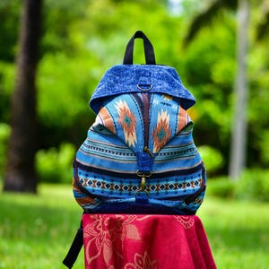 Op de afbeelding: Een blauwe rugzak met een op het zuidwesten geïnspireerd patroon. De tas heeft een klepsluiting, een handvat aan de bovenkant en verstelbare bandjes. Het ontwerp bevat tinten blauw, bruin en oranje, met een decoratief patroon. De rugzak staat op een rode doek.