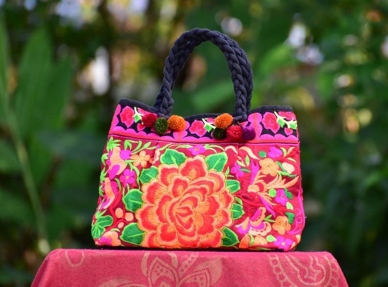 May include: A pink and red embroidered tote bag with black handles and colorful pom-poms. The bag features a large floral design with a central rose.