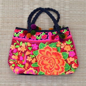 May include: A pink and red floral tote bag with black braided handles and colorful pom-poms. The bag is embroidered with a large floral design.