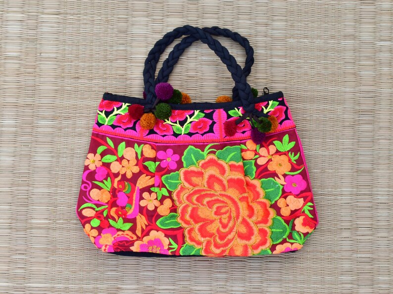 May include: A pink and red floral tote bag with black braided handles and colorful pom-poms. The bag is embroidered with a large floral design.