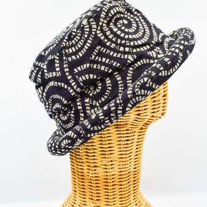 Op de afbeelding: Een bucket hat van donkerblauwe stof met een herhalend, gebroken wit spiraalpatroon. De hoed heeft een ronde kroon en een naar beneden lopende rand die het hoofd omsluit, met een consistent textielpatroon over het gehele ontwerp.