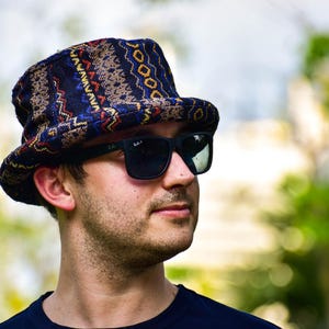 Könnte beinhalten: Ein gemusterter Fedora-Hut mit dunkelblauer Basis und einem geometrischen Design in Rot, Gelb und Weiß. Die Person trägt eine schwarze Sonnenbrille und ein dunkelblaues Oberteil. Der Hintergrund ist mit grünem Laub verschwommen.