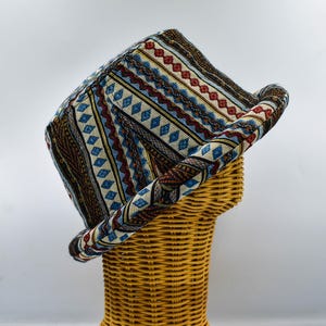 May include: A woven hat with a rectangular crown and a rolled brim. The hat features a colorful pattern of blue, red, yellow, and brown geometric designs. The hat is displayed on a wicker mannequin head.