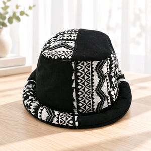 Può includere: Un cappello a secchiello con un design patchwork composto da pannelli neri a tinta unita e pannelli bianchi con un motivo geometrico nero. Il cappello presenta una corona arrotondata e una tesa larga inclinata verso il basso con il motivo.
