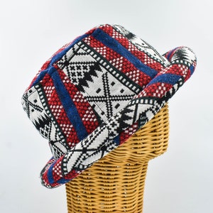 Può includere: Cappello fantasia con parte superiore piatta e bordo arrotolato. Il cappello presenta un motivo geometrico in rosso, blu, nero e bianco. Il cappello è esposto su una forma di testa in vimini su sfondo bianco.