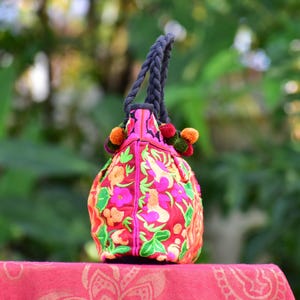 May include: A pink tote bag with colorful floral embroidery and black rope handles. The bag has pom-poms in pink, green, and orange.