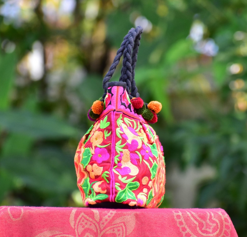 May include: A pink tote bag with colorful floral embroidery and black rope handles. The bag has pom-poms in pink, green, and orange.