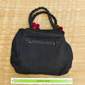 May include: Black canvas tote bag with a zippered pocket and a braided handle. The bag is decorated with colorful fabric flowers.