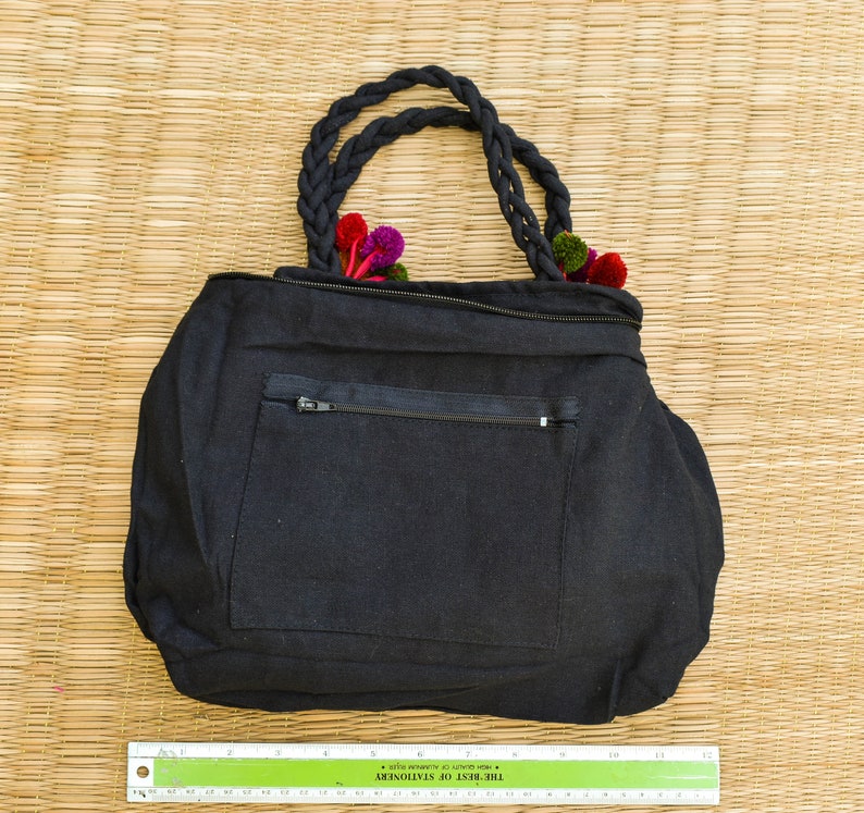 May include: Black canvas tote bag with a zippered pocket and a braided handle. The bag is decorated with colorful fabric flowers.