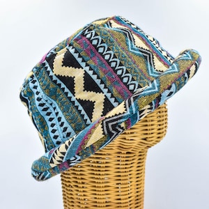 Può includere: Cappello fantasia con tesa, caratterizzato da un design geometrico in verde acqua, nero, beige e viola. Il cappello è esposto su una forma di vimini, che ne mostra la forma e la consistenza. Il design del cappello include zigzag e altre forme geometriche.