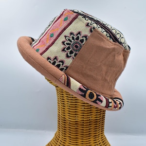 May include: A fedora hat with a patchwork design. The hat has a brown brim and a crown with patterned fabric in pink, beige, and black. The hat is displayed on a wicker stand.