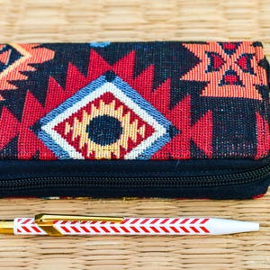 May include: A rectangular black wallet with a colorful geometric pattern in red, orange, and blue. A white pen with red chevron stripes and gold accents lies below the wallet. A green pom-pom hangs from the zipper.