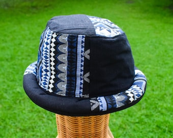 May include: A bucket hat featuring a patchwork design of solid black fabric panels and panels with a blue, white, and black geometric woven pattern. The hat has a rounded crown and a wide, downward-sloping brim that matches the patchwork style.
