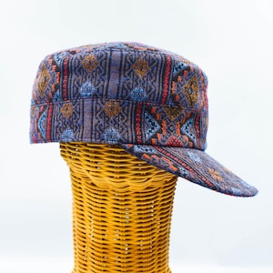May include: A military-style cap featuring a woven fabric with a dark purple base and a geometric pattern in shades of blue, orange, and red. The hat has a flat top, a structured crown, and a stiff, extended brim with the same intricate pattern.