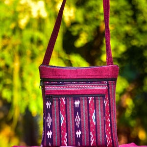 Pode incluir: Bolsa transversal retangular com um design estampado. A bolsa apresenta uma cor magenta escura com listras verticais em vermelho, verde-azulado e branco. Possui um zíper na parte superior e uma alça longa e fina. A bolsa é feita de tecido.