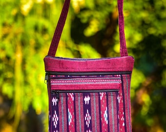 May include: A rectangular crossbody bag with a patterned design. The bag features a deep magenta color with vertical stripes in red, teal, and white. It has a zippered top and a long, thin strap. The bag is made of fabric.