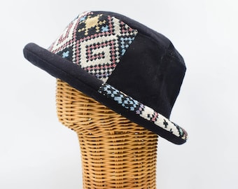 May include: A black bucket hat featuring a woven panel with a geometric pattern in cream, red, and blue. The hat has a solid black crown and a matching black brim with a decorative woven trim along the edge, creating a distinct contrast.