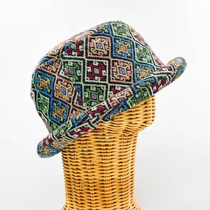 May include: A colorful fedora hat with a geometric pattern in shades of blue, green, red, and yellow. The hat is displayed on a wicker head form against a white background. The hat's brim is slightly curved.