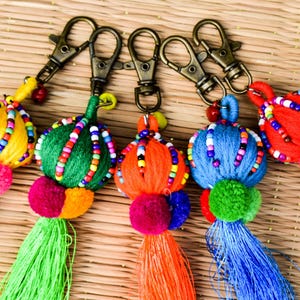 Op de afbeelding: Een verzameling kleurrijke sleutelhangers met metalen sluitingen. Elke sleutelhanger heeft een met garen omwikkelde bol versierd met kralen, een pompon en een kwast. De sleutelhangers zijn geel, groen, oranje, blauw en rood.