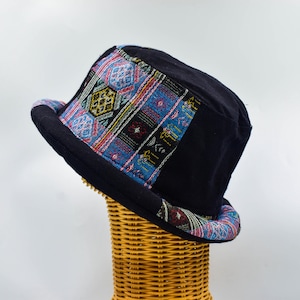 Op de afbeelding: Zwarte bucket hat met een kleurrijke patroonband. De band heeft geometrische ontwerpen in blauw, rood, geel en groen. De hoed wordt gepresenteerd op een geweven rieten standaard tegen een witte achtergrond.