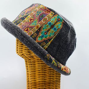 Pode incluir: Um chapéu de feltro cinza-escuro com uma faixa larga de tecido colorido com padrões geométricos complexos em tons de turquesa, vermelho, amarelo e preto. O chapéu tem uma copa estruturada e uma aba ligeiramente levantada.
