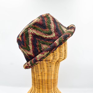 May include: A patterned hat with a brim, featuring a woven design in red, green, and navy blue. The hat is displayed on a wicker head form against a white background. This hat is a stylish accessory. The hat's design is reminiscent of a tapestry.