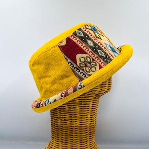 Könnte beinhalten: Ein gelber Fedora-Hut mit einem gemusterten Band und Seitenteil. Das Band weist ein farbenfrohes geometrisches Design mit roten, blauen und cremefarbenen Akzenten auf. Der Hut wird auf einem Weiden-Mannequinkopf präsentiert.