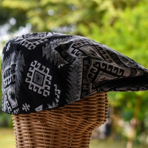 May include: A black and gray patterned flat cap. The cap features a repeating geometric design in shades of gray and black. The cap is displayed on a wicker mannequin head against a blurred green background.